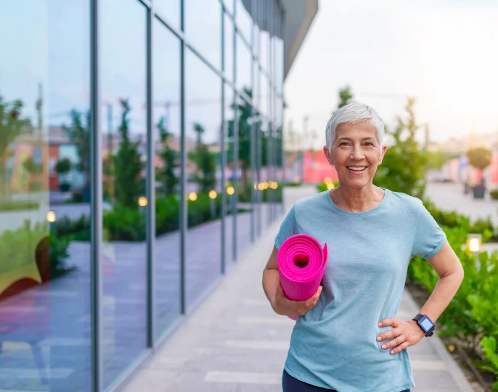 happy senior woman walking on the sidewalk holding yoga mat